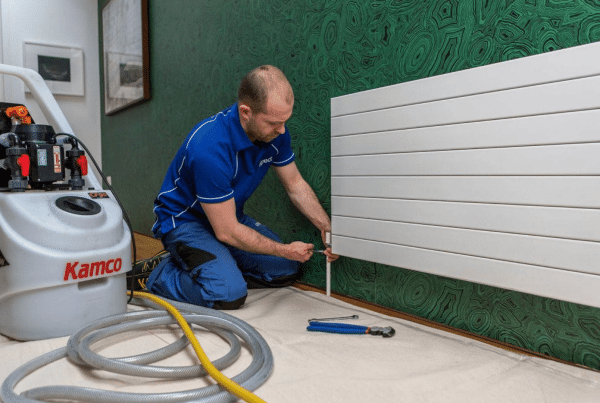 A technician in a blue uniform kneels on a mat, using tools to work on a white radiator attached to a green patterned wall, with a Kamco powerflushing machine and hoses placed nearby.