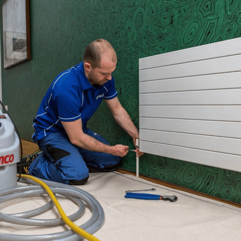 A technician in a blue uniform kneels on a mat, using tools to work on a white radiator attached to a green patterned wall, with a Kamco powerflushing machine and hoses placed nearby.