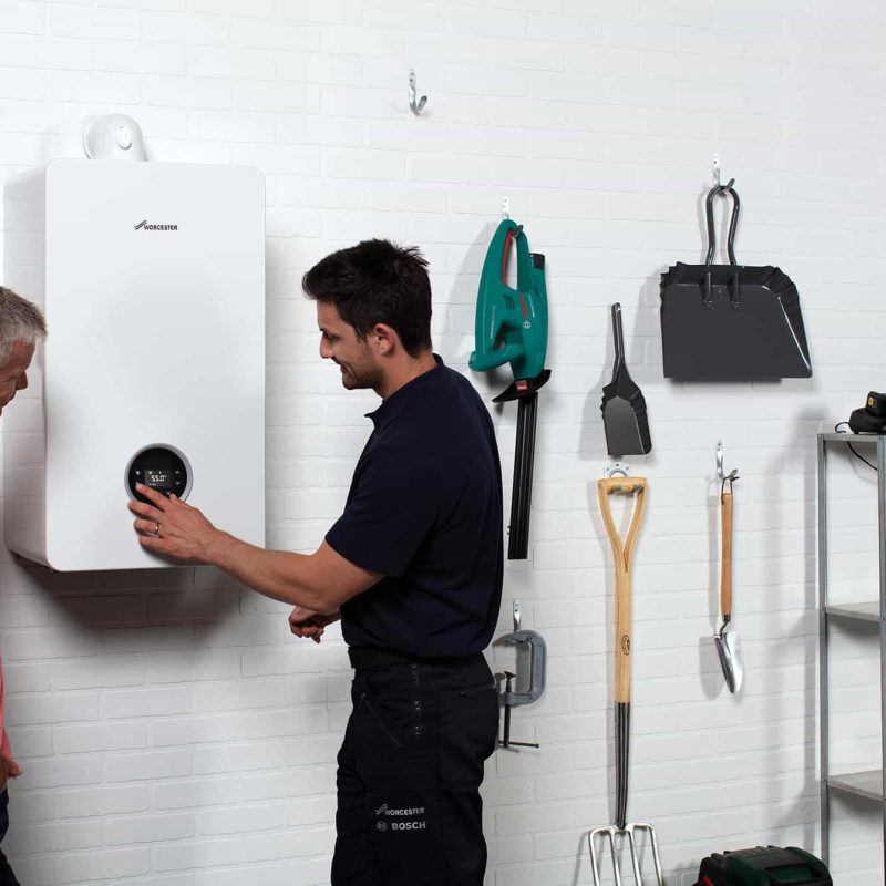 A technician discusses a Worcester Bosch wall-mounted boiler with a man in a garage, surrounded by tools and gardening equipment hanging on the white brick wall—demonstrating expert boiler installation.