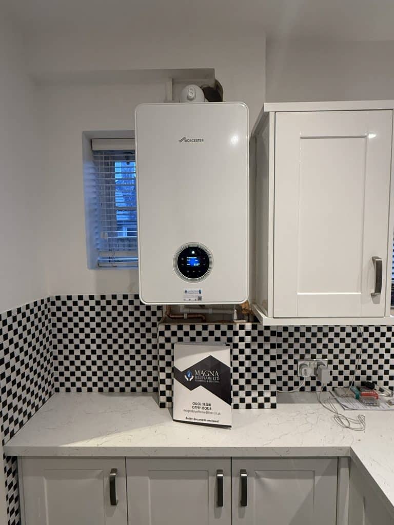 A white wall-mounted boiler with a digital display is installed above a kitchen worktop with black-and-white chequered tiles. A Magna box and some kitchen cupboards are also visible.
